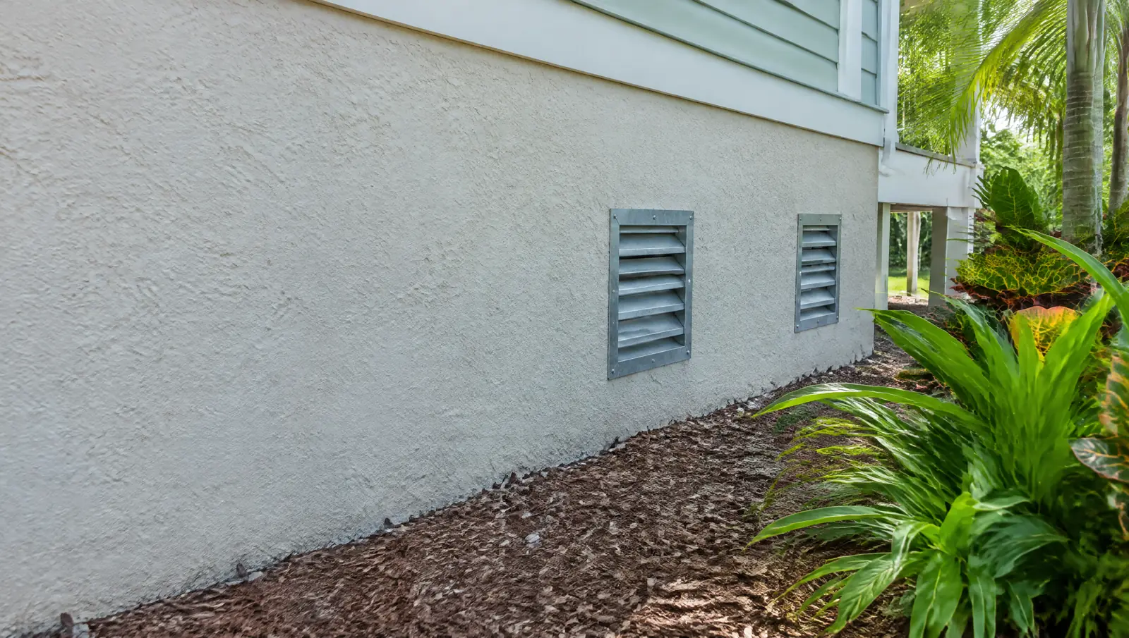 Two louvred metal flood vents in the stucco foundation wall of a raised Florida home