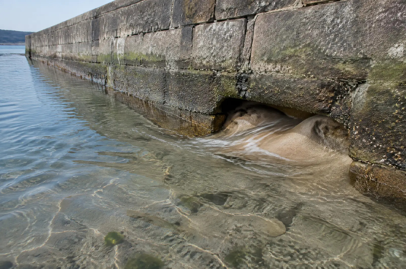 Gap at the base of an old seawall at low tide with sand washing out from behind the wall